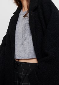 Czarny puszysty płaszcz nałożony na jasnoszary prążkowany krótki sweter, zestawiony z czarnymi kratkowymi spodniami z guzikami.