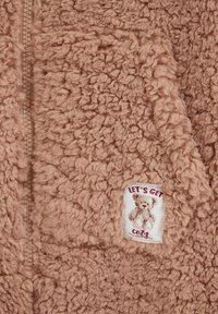 Veste polaire teddy marron douce avec une fermeture éclair visible et un patch montrant un ours en peluche et le texte "LET'S GET COZY."