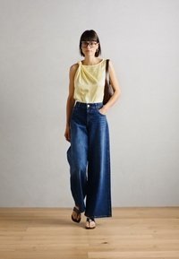 Top sem mangas amarelo claro com detalhe drapeado, combinado com jeans de denim azul escuro de perna larga, usado com sandálias pretas. Fundo simples.