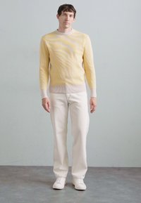 Gelb-weißer gestreifter Pullover mit strukturiertem Rippenmuster, kombiniert mit weißen Jeans und weißen Sneakers, vor einem hellen Hintergrund stehend.