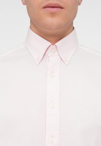 Camisa de botón de color rosa claro con un cuello clásico, cinco botones en el frente y una tela suave y texturizada.