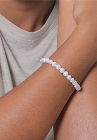Personne portant un bracelet de perles blanches au poignet, vêtue d'un haut gris clair, les bras croisés.