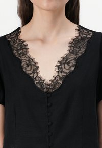 Haut noir à manches courtes, avec un col en V orné d'une bordure en dentelle noire et de boutons décoratifs sur le devant.