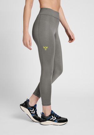 Woman wearing high-waisted gray leggings with yellow bee logo on thigh and black-blue athletic shoes, standing sideways on gray floor.