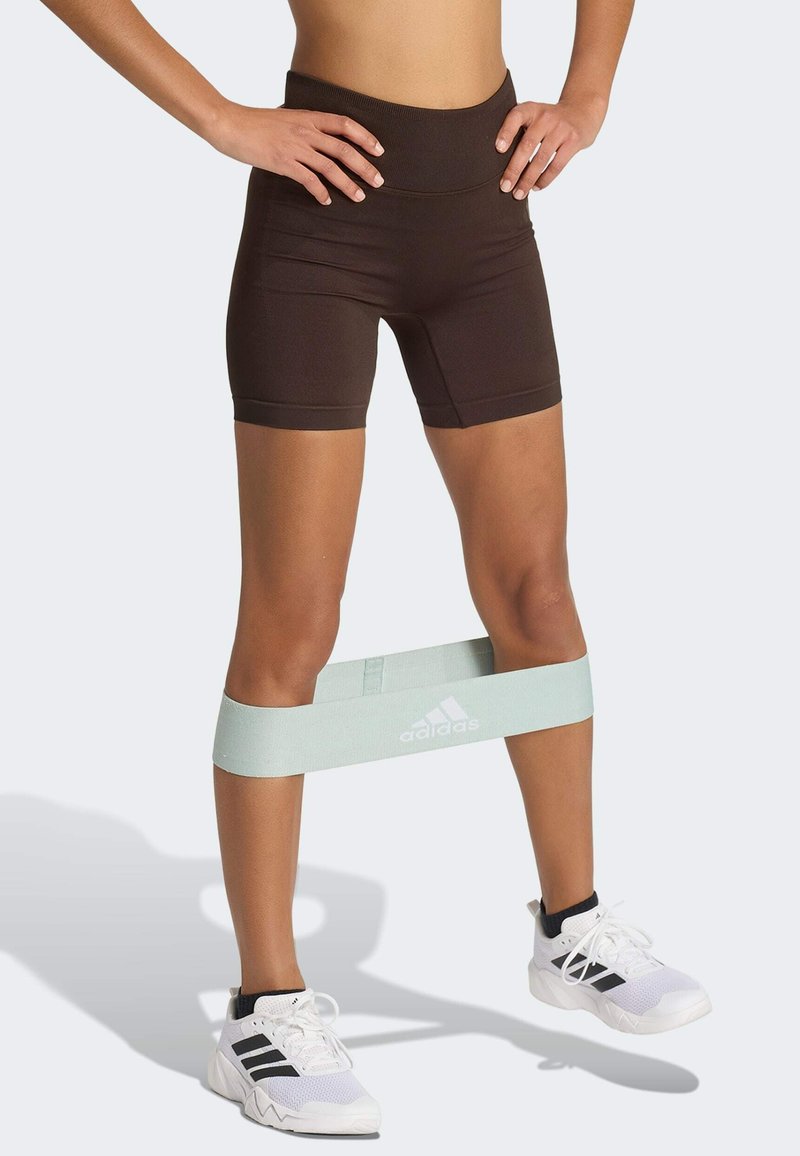 Piernas con pantalones cortos negros y zapatillas blancas Adidas con una banda de resistencia verde claro alrededor de las piernas inferiores, manos apoyadas en las caderas.