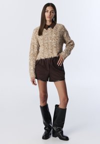 Mujer vistiendo un suéter beige texturizado sobre una camisa marrón con cuello, pantalones cortos marrones oscuros y botas de cuero negras hasta la rodilla, de pie contra un fondo blanco.