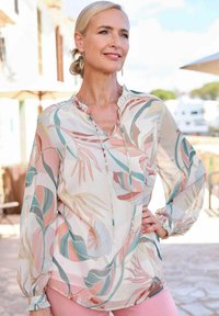 Bluse mit Blumenmuster aus leichtem, pastellfarbenem Stoff, verfügt über lange Ärmel, einen gerafften Ausschnitt und tropische Blätter in Rosa-, Grün- und Blautönen.