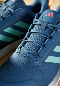 Scarpe da ginnastica blu con una parte superiore testurizzata, sezioni in rete e strisce blu chiaro. Presentano un logo rosa sulla linguetta e lacci piatti.