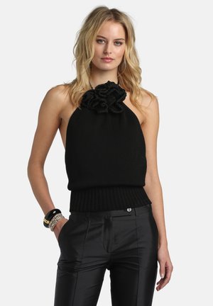 Žena u crnom topu bez rukava s volančićima na izrezu i crnim satenskim hlačama, ukrašena narukvicama na lijevom zapešću.