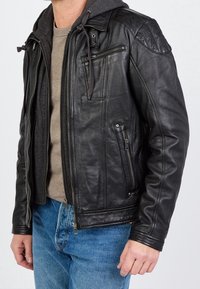 Zwarte leren jas met capuchon, voorzien van ritszakken, een gladde textuur en subtiele stikseldetails. Gedragen over een grijs shirt en blauwe spijkerbroek.