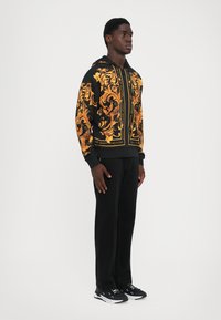 Homme se tenant debout les mains sur les côtés, portant un pantalon noir, un hoodie noir et doré à motif, et des baskets noir et blanc sur fond blanc.