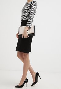 Haut à manches longues à motif zigzag noir et blanc, jupe crayon noire, escarpins en vernis noir et une pochette rayée beige et noire.