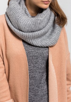 Femme portant un manteau rose clair, un pull gris foncé et une écharpe infinity en grosse maille gris clair autour du cou.