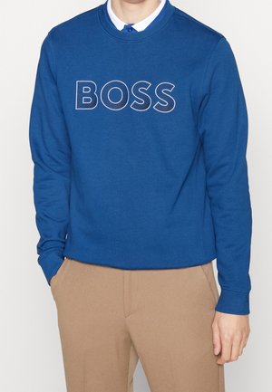 Mann trägt einen blauen Sweatshirt mit langen Ärmeln und dem Schriftzug "BOSS", beige Hose und ein weißes Hemd darunter, steht mit den Händen in den Taschen.