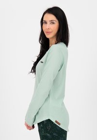 Mintgroene longsleeve top van zacht materiaal, met een ronde hals en zijsplitten. Het ontwerp bevat een bruin logo-label aan de onderrand.