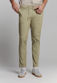 Khaki stretchbroek met een elastische taille, tapered fit en zakken, gecombineerd met grijze sportschoenen.