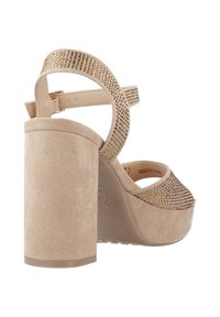 Sandal de tacón alto beige con plataforma de ante redondeada, correa decorada con tachuelas doradas y correa ajustable en el tobillo para un ajuste seguro.