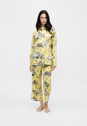 Une femme porte un ensemble de pyjama jaune boutonné, imprimé de motifs d’animaux en noir et blanc. Elle se tient pieds nus devant un fond clair uni.