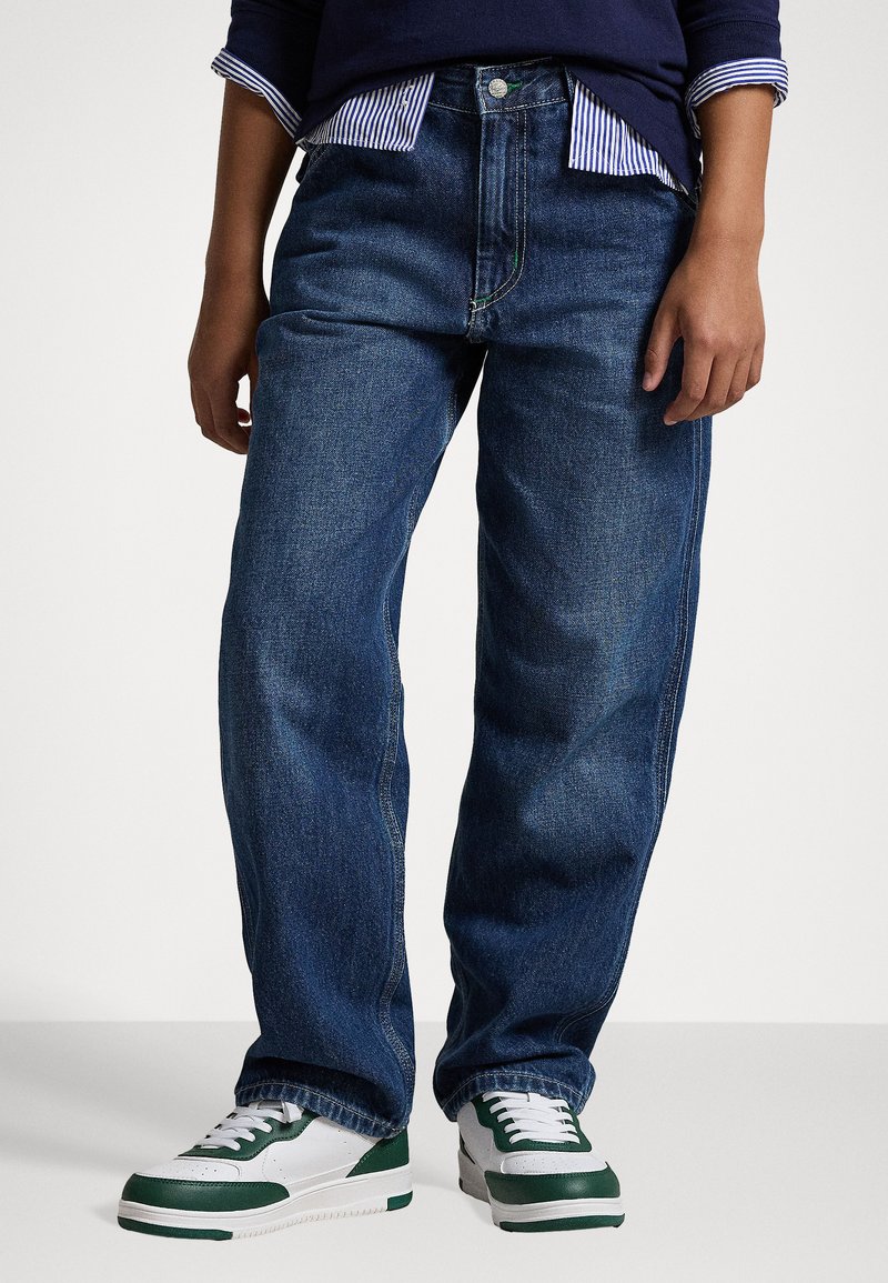 Jeans de ganga azul escuro com um design de perna reta, apresentando um fecho de botão e detalhes de costura visíveis. Combinados com sapatilhas brancas e verdes.