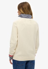 Crèmewitte gebreide trui met een geribde zoom en gestreepte kraag. Accenten van rode en marineblauwe strepen op de mouwen. Casual ontwerp.