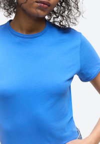 Femme aux cheveux bouclés portant un t-shirt bleu vif à manches courtes et col rond, posant avec la main sur la hanche devant un fond blanc.