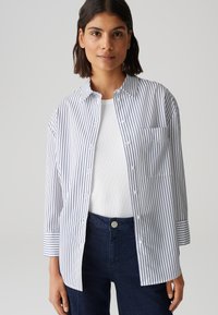 Camicia bianca con righe verticali navy, maniche corte e una sola tasca sul petto, abbinata a jeans blu scuro. Tessuto dalla trama liscia.