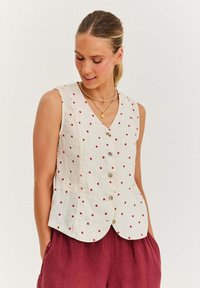 Biała lniana kamizelka w czerwone kropki, z dekoltem w serek, na guziki z przodu i dwoma kieszeniami z przodu. Bezrękawnik, połączony z luźnymi bordowymi spodniami.