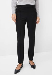 Un pantalon noir ajusté présente une coupe slim, des poches zippées sur les côtés et une texture lisse. Porté avec des ballerines noires à bout pointu.