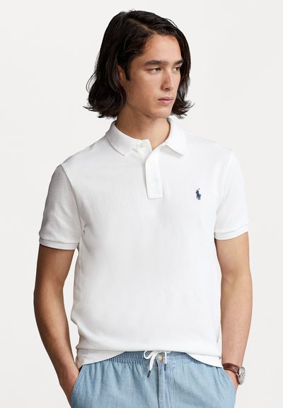 Hvit polo-skjorte laget av bomull, med klassisk krage, korte ermer og en liten brodert logo i blått på brystet.