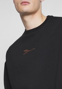 Primo piano di una persona che indossa una maglietta nera a girocollo con un piccolo ricamo arancione della firma sul petto.