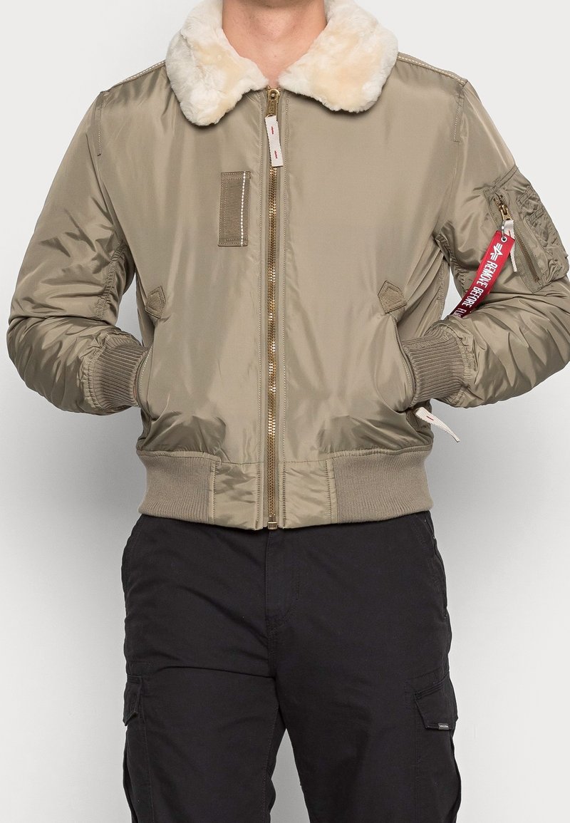 Giacca da volo in nylon taupe con colletto in pelliccia sintetica. Presenta polsini e orlo a costine, zip frontale e una tasca sulla manica con etichetta rossa.