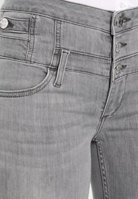 Los jeans de mezclilla gris presentan un diseño de cintura alta, estructura de cinco bolsillos y cierre de tres botones. El material tiene una textura suave y costuras sutiles.