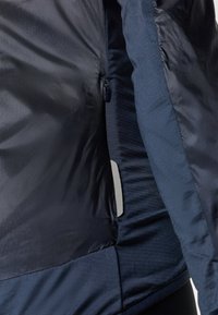 Mornarice plavi kaput s zatvaračem na zip, glatkom vanjskom tkaninom i teksturiranom unutarnjom podstavom. Uključuje bočnu džep s srebrnim akcentom.