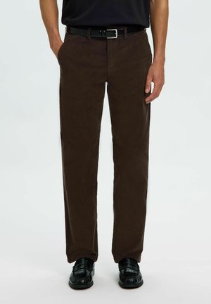 Homme portant un pantalon marron foncé droit, ceinture noire, chemise noire et mocassins noirs, debout les mains détendues.