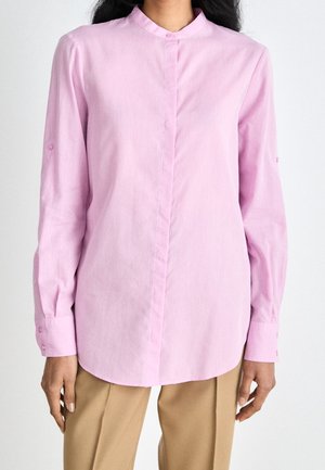 Žena v světle růžové košili s dlouhým rukávem a mandarínským límečkem a béžových kalhotách, stojící před jednobarevným pozadím.