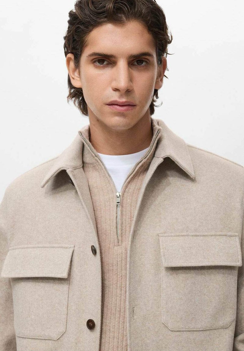 Giacca in misto lana beige con colletto, due tasche sul petto e bottoni color legno. Indossata sopra un top in maglia beige con zip.