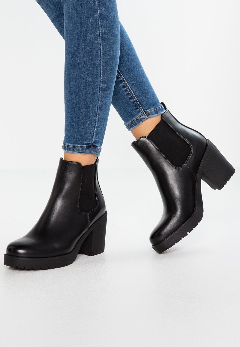 Musta nahkasaapas nilkkapituudella, jossa on paksu korko, joustavat sivupaneelit ja teksturoitu kumipohja, yhdistettynä hyvin istuviin sinisiin farkkuihin.