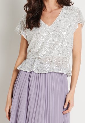 Vrouw die een wit pailletten top met korte mouwen en een V-hals draagt, gecombineerd met een lichtpaarse geplooide rok, staand tegen een effen achtergrond.