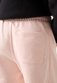 Un pantalon de survêtement rose clair avec une poche arrière, une taille élastique et une texture de tissu lisse, avec un haut noir visible au-dessus.