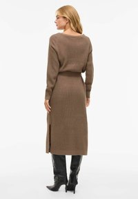 Długa, dzianinowa sukienka w ziemistym brązie z teksturowanym wzorem, z wysokim rozcięciem po lewej stronie i dopasowaną talią. W zestawieniu z czarnymi kozakami za kolano.