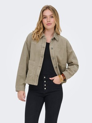 Ung kvinde med skulderlangt blondt hår iført en beige kordfløjlsjakke over en sort top og sorte højhæsede bukser med knapper.