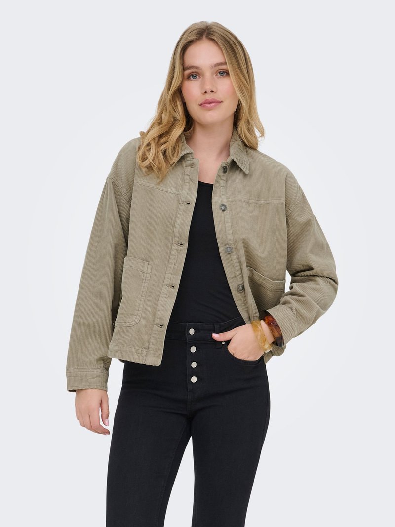 Jeune femme aux cheveux blonds mi-longs, portant une veste en velours côtelé beige sur un haut noir et un pantalon noir taille haute avec des boutons.