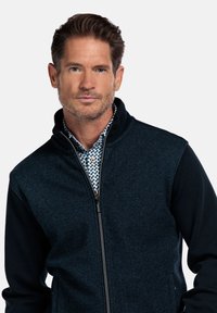 Marinefarbende Zip-Jacke mit strukturiertem Vorderteil und einfarbigen dunklen Ärmeln. Sie hat einen hohen Kragen und steht vor einem einfarbigen Hintergrund.