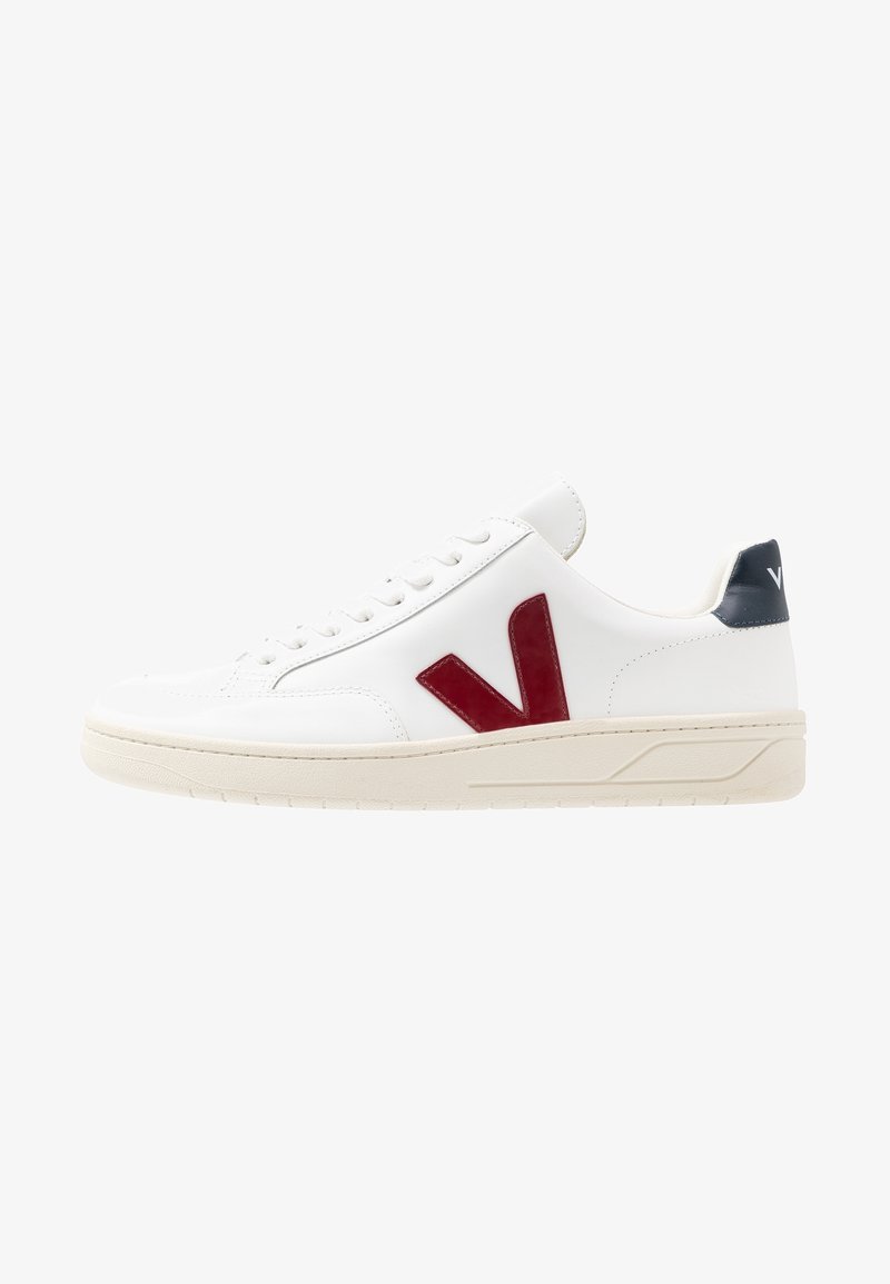 Baskets basses blanches avec logo rouge "V" sur le côté, lacets blancs, semelle beige et languette arrière noire avec inscriptions blanches.