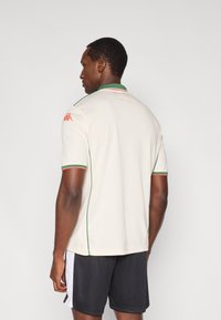 Kreminės spalvos trumpomis rankovėmis marškinėliai su žaliais ir raudonais akcentais; pasižymi ribbine tekstūra, raudonu Kappa logotipu ant kairiojo peties ir kontrastine apykakle.