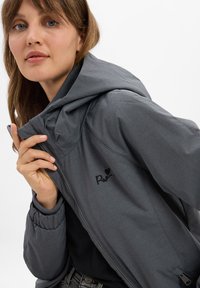 Graue Kapuzenjacke mit einer glatten Oberfläche, Reißverschluss, gerippten Bündchen und einem bestickten Logo. Modell hält die Jacke teilweise offen.