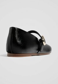 Ballerina nera in pelle con punta arrotondata, caratterizzata da una fibbia decorativa dorata e una suola bassa marrone, texture liscia e design minimalista.
