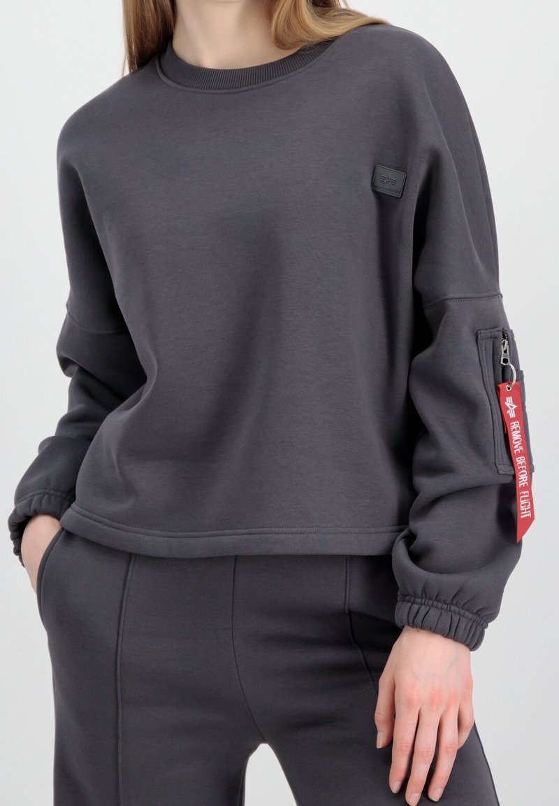 Sweatshirt gris foncé en tissu doux, avec un col rond, des poignets élastiques et une poche zippée sur le côté avec un détail d'étiquette rouge.