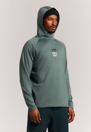 Man die een groen hooded shirt met lange mouwen draagt met het "PRO TEST" logo en een zwarte gebreide muts, staand tegen een effen lichte achtergrond.