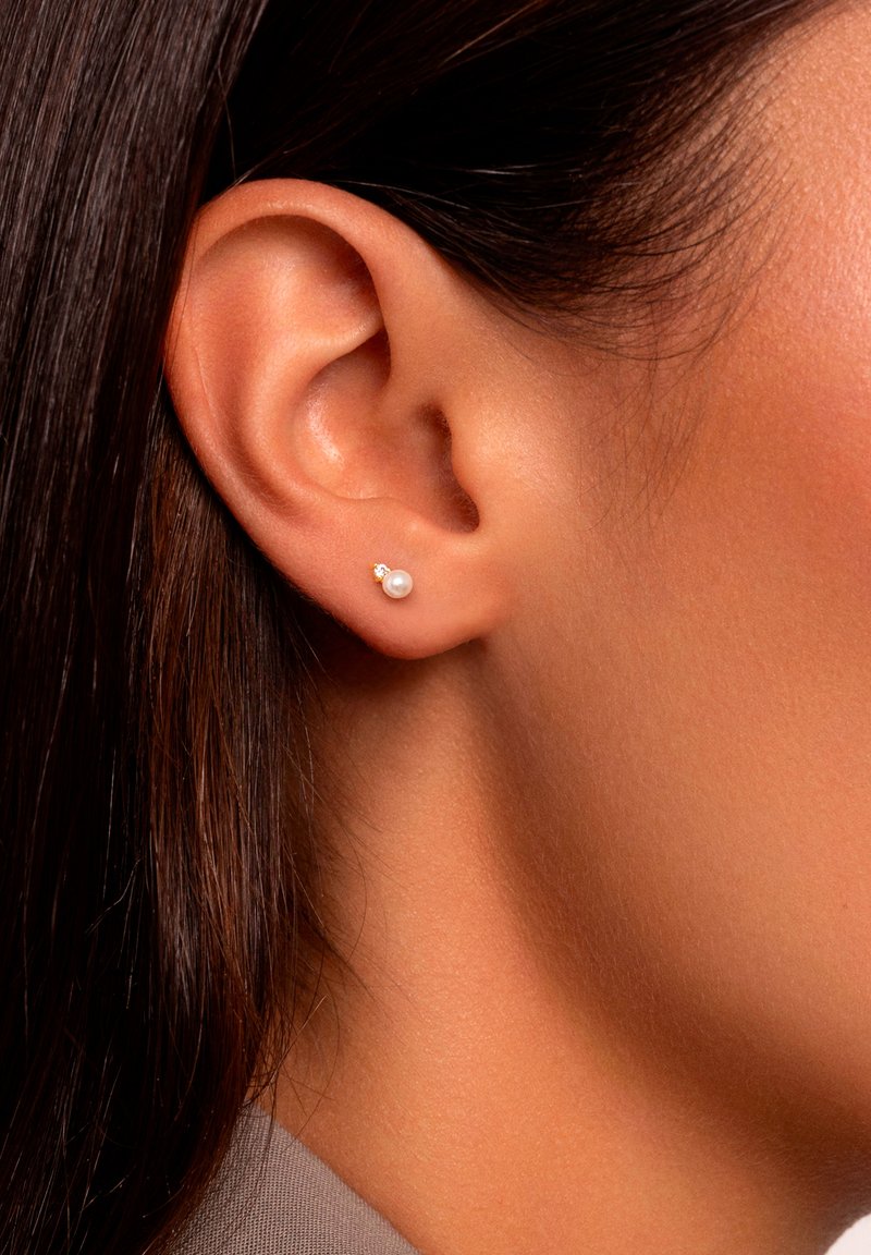 Pendientes de perno de oro que presentan una pequeña perla blanca y un pequeño acento de gema clara, sobre piel suave y bronceada y cabello negro lacio.